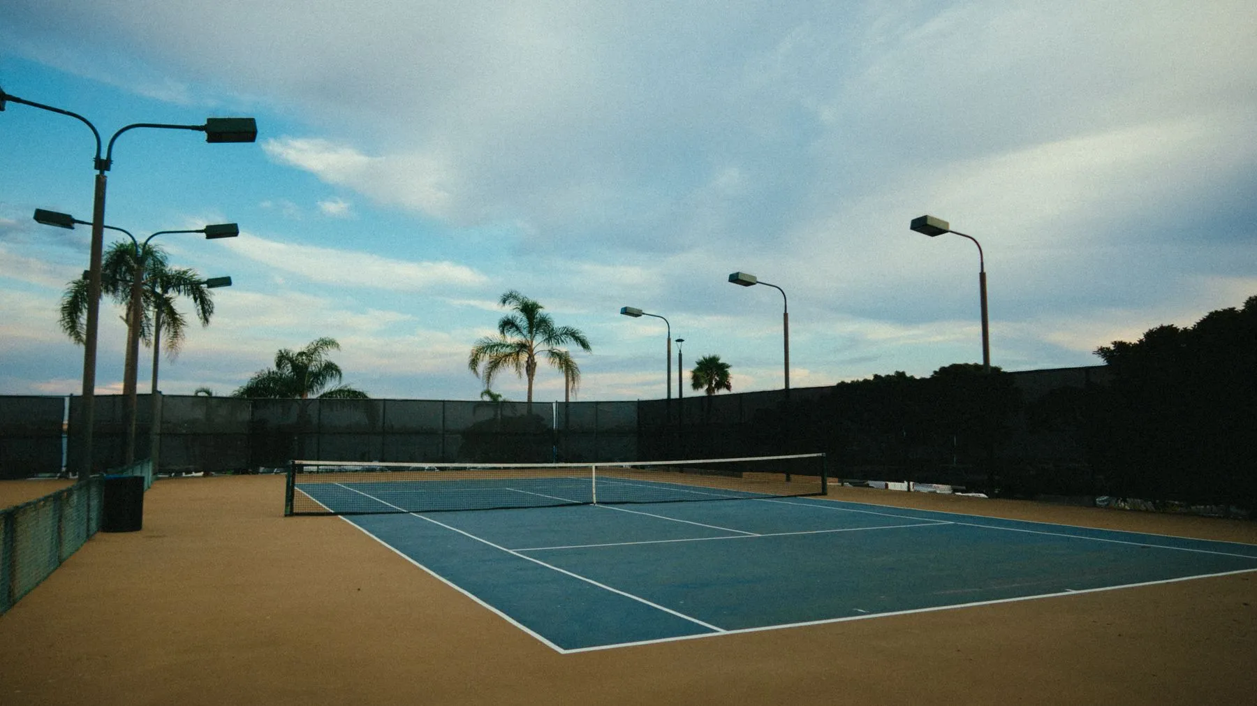 Cancha de tenis al atardecer, con palmeras al fondo.